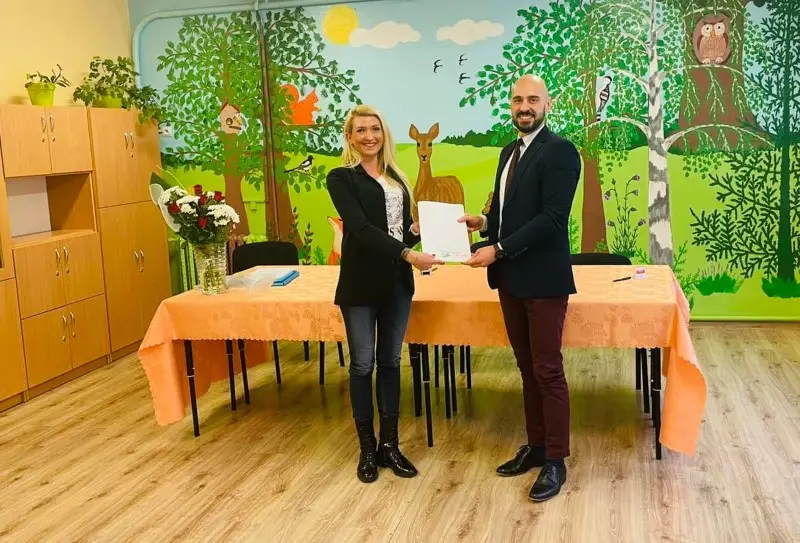 Zdjęcie wykonane w orientacji poziomej przedstawia kobietę i mężczyznę stojących w sali przedszkolnej przed stołem przykrytym pomarańczowym obrusem. Kobieta po lewej stronie ma na sobie czarną marynarkę i ciemne jeansy, natomiast mężczyzna po prawej jest ubrany w ciemną marynarkę, białą koszulę i bordowe spodnie. Razem trzymają biały dokument, który jest centralnym punktem kompozycji, podkreślając charakter oficjalnego spotkania lub podpisania umowy. Tło stanowi kolorowy mural z motywem leśnym, przedstawiający drzewa, sarnę, sowę oraz inne elementy przyrody typowe dla edukacji wczesnoszkolnej. Po lewej stronie kadru widać jasne drewniane szafki oraz wazon z bukietem róż, co dopełnia wystrój wnętrza o przytulnym, profesjonalnym charakterze.