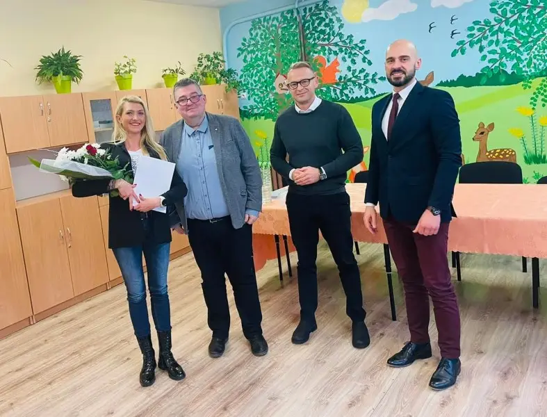 Zdjęcie wykonane w orientacji poziomej przedstawia grupę czterech osób stojących w jasnej sali przedszkolnej. Po lewej stronie znajduje się kobieta w czarnym żakiecie i jeansach, trzymająca bukiet kwiatów oraz białą teczkę, a obok niej stoi trzech mężczyzn w strojach półformalnych i garniturowych. W tle widać meble w kolorze jasnego drewna oraz ścianę z namalowanym kolorowym krajobrazem przedstawiającym drzewa, zwierzęta i słońce. Na pierwszym planie po prawej stronie widoczny jest stół przykryty brzoskwiniowym obrusem, przy którym ustawiono czarne krzesła. Całość kompozycji utrzymana jest w jasnej, przyjaznej tonacji, sugerującej spotkanie w placówce edukacyjnej lub kulturalnej. Oświetlenie jest równomierne, dobrze podkreślające detale wnętrza oraz uśmiechnięte twarze uczestników spotkania.