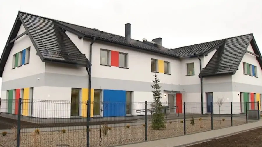 Zdjęcie przedstawia nowoczesny budynek użyteczności publicznej w formacie poziomym o proporcjach zbliżonych do 16:9. Dwupiętrowa konstrukcja wyróżnia się białą elewacją z szarymi pasami oraz licznymi, kolorowymi akcentami w odcieniach czerwieni, żółci, niebieskiego i zieleni, umieszczonymi przy oknach. Budynek posiada dwuspadowy dach pokryty ciemną dachówką oraz system rynnowy w ciemnym kolorze. Teren wokół obiektu jest ogrodzony metalowym panelem, a przed wejściem znajduje się podjazd dla osób niepełnosprawnych oraz niewielkie nasadzenia iglaków. Całość sprawia wrażenie przyjaznej i nowoczesnej placówki edukacyjnej lub przedszkola, utrzymanej w jasnej, uporządkowanej kolorystyce.